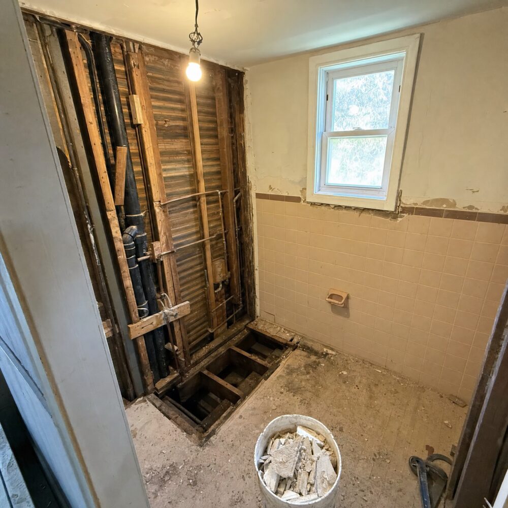 A residential bathroom mid-demolition with tile stripped and the subfloor opened up