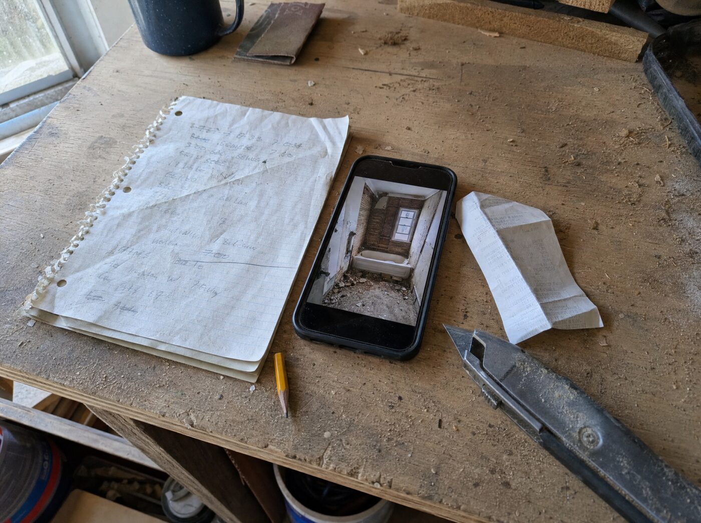 A workbench with a messy notepad, a phone showing a job-site photo and a supplier receipt